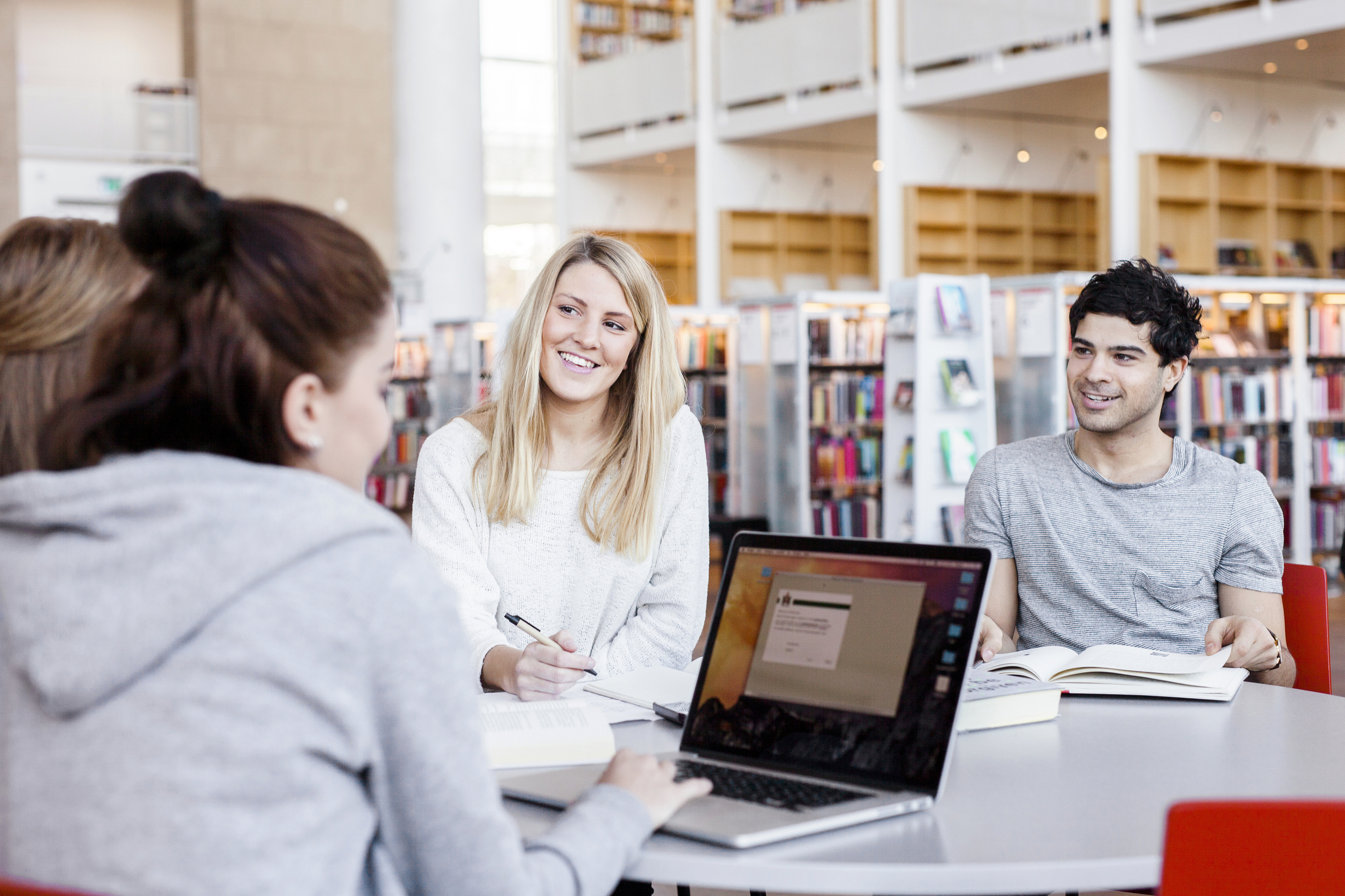 scandinav_evkb Tre unga vuxna sitter runt ett bord med en dator igång. I bakgrunden syns böcker i ett bibliotek. Foto: Astrakan Images AB/Scandinav