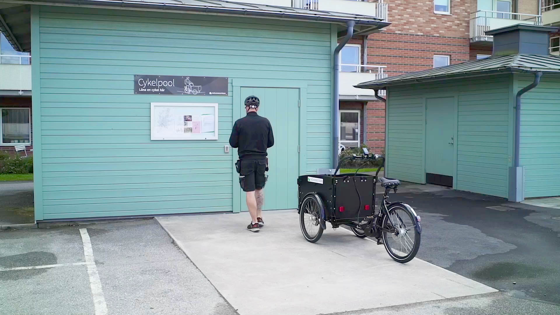 cykelpool-byggnad Man är påväg mot ett litet hus där cykelpoolen finns. En cykel med flak står bredvid.